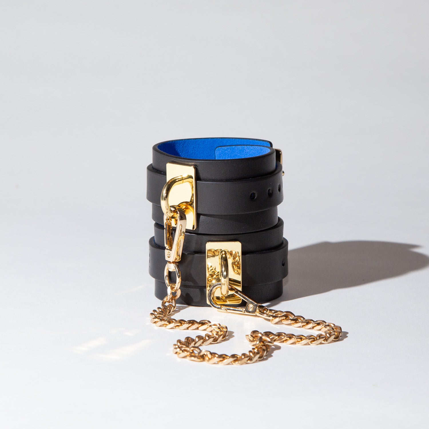 Black leather wrist cuffs with gold chain and blue suede interior on a light gray background