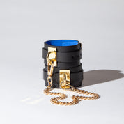 Black leather wrist cuffs with gold chain and blue suede interior on a light gray background